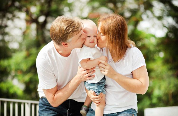 toddler-photoshoot-tips-parents-kissing-studio Toddler with parents kissing her cheeks during photoshoot